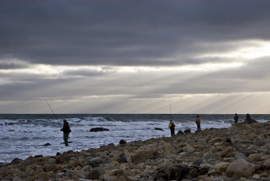A man casts a fishing line off of a rocky northeastern beach under cloudy skies.
