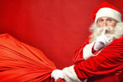 Santa Claus holding a bag of toys and putting his index finger to his lips in a shushing motion.