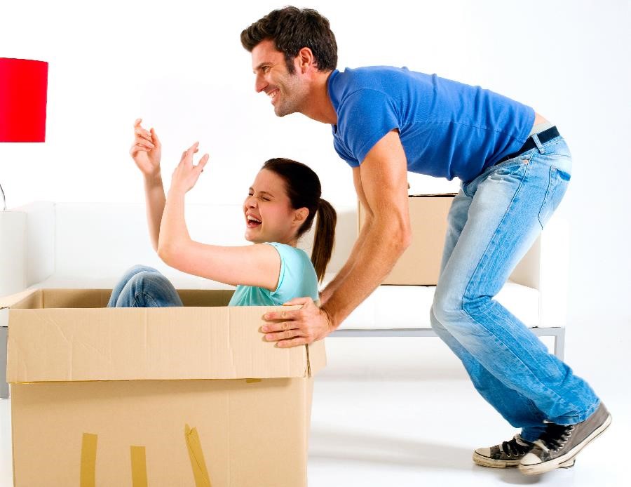 A smiling man pushing a moving box while a laughing woman sits in it.