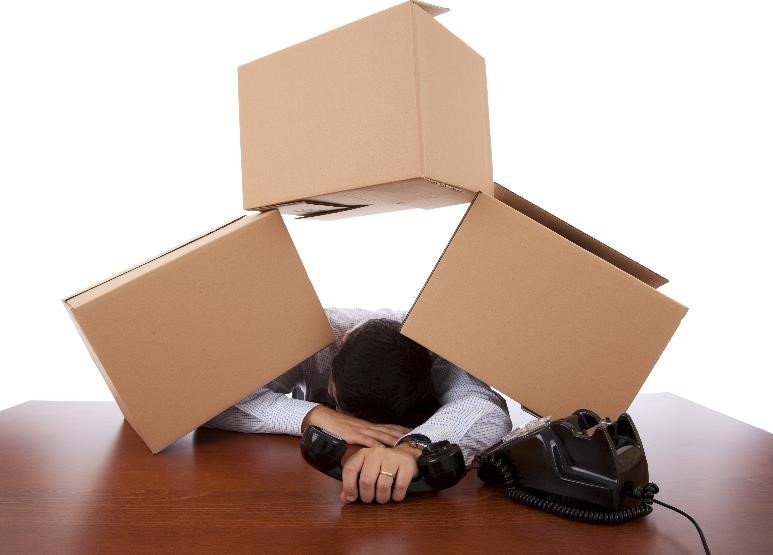 A businessman with three boxes sitting on top of him while he rests his head against his desk and holds a phone receiver.