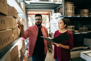 A man pointing at boxes on shelves talking to a woman with a folder.